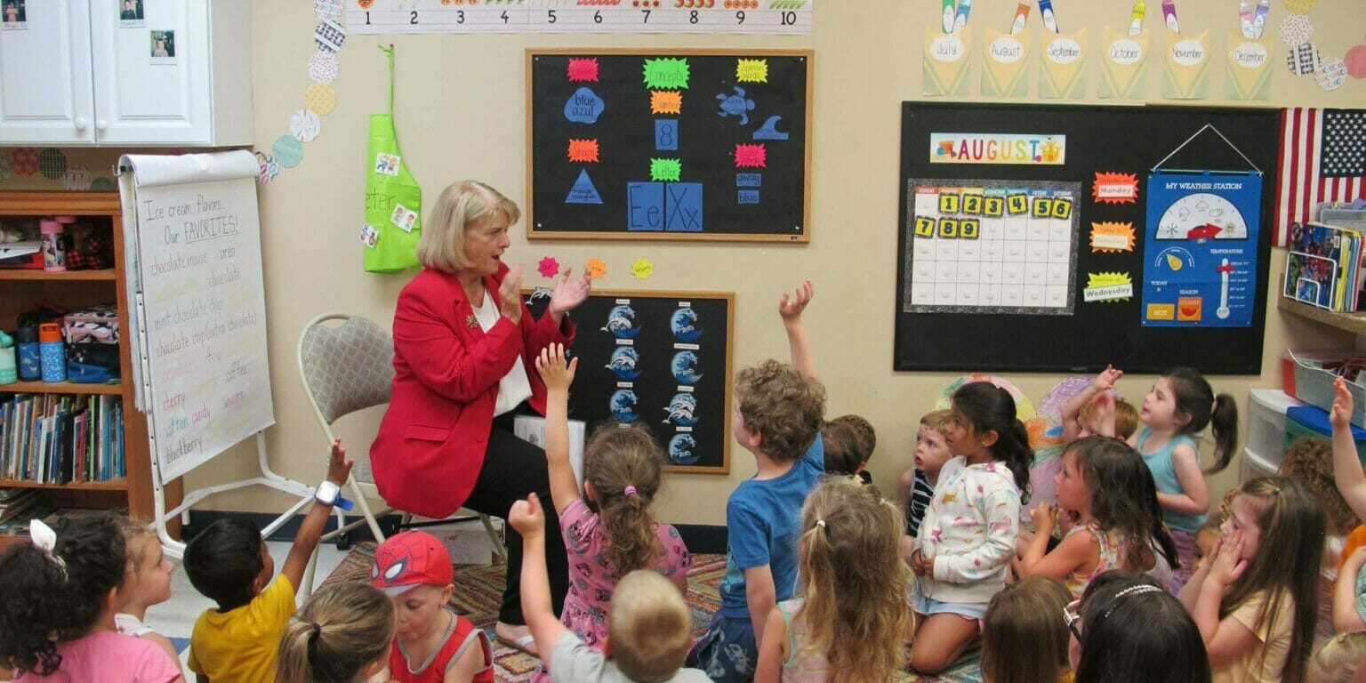 mary-ann-answering-questions-cmwm-preschool-smithfield-8-9-22 Woman in red jacket answers questions from large group of children with their hands raised.